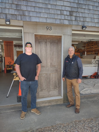 Two men stand smiling in front of a wooden door marked